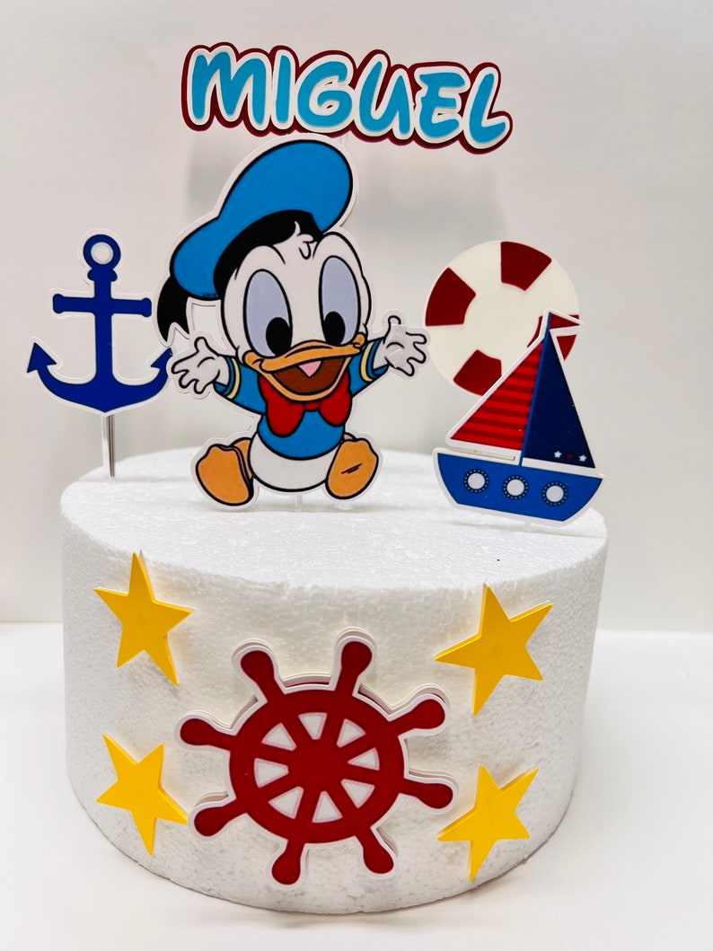 May include: A white cake decorated with a blue and yellow nautical themed cake topper. The topper features a cartoon duck, an anchor, a life preserver, a sailboat, and a red steering wheel. The cake is decorated with yellow stars. The cake topper says "Miguel".