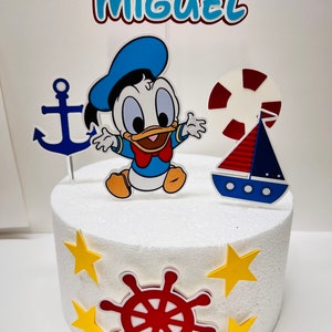 May include: A white cake decorated with a blue and yellow nautical themed cake topper. The topper features a cartoon duck, an anchor, a life preserver, a sailboat, and a red steering wheel. The cake is decorated with yellow stars. The cake topper also includes the name "MIGUEL".