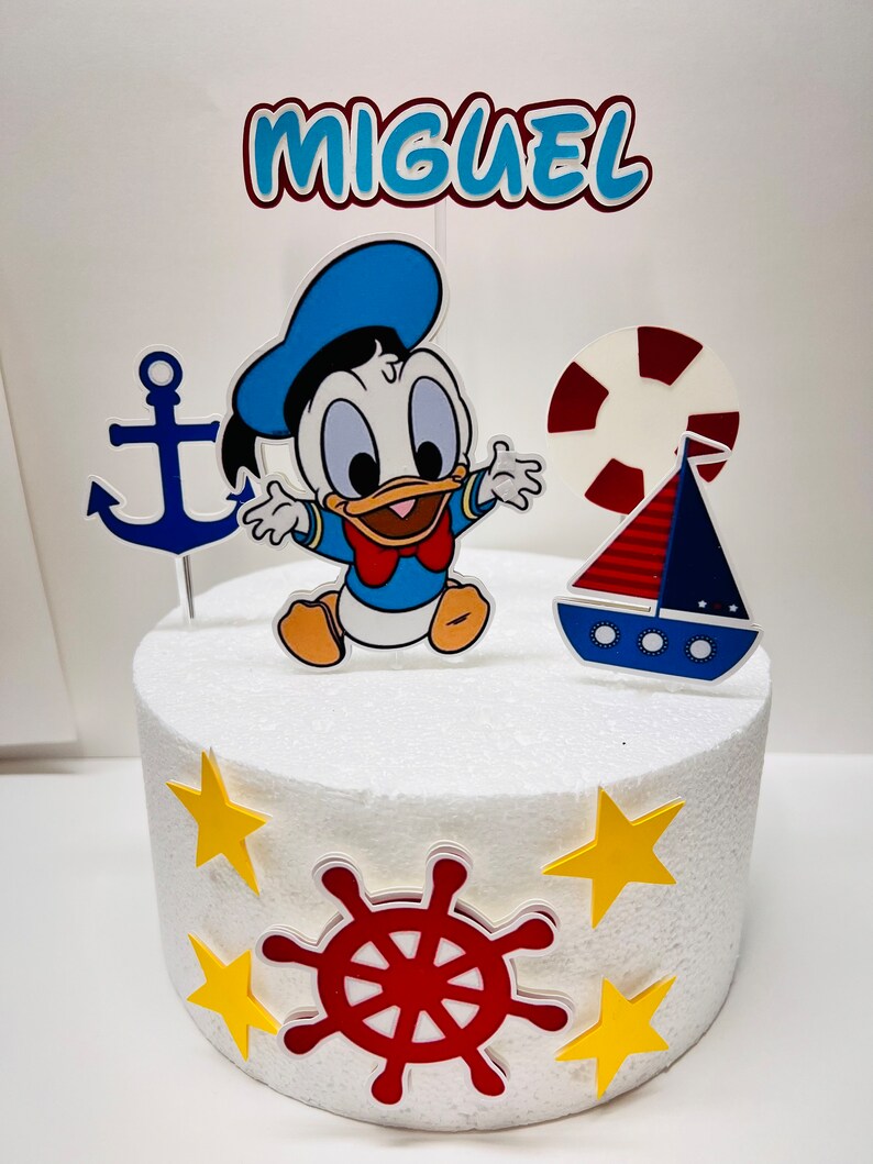 May include: A white cake decorated with a blue and yellow nautical themed cake topper. The topper features a cartoon duck, an anchor, a life preserver, a sailboat, and a red steering wheel. The cake is decorated with yellow stars. The cake topper also includes the name "MIGUEL".