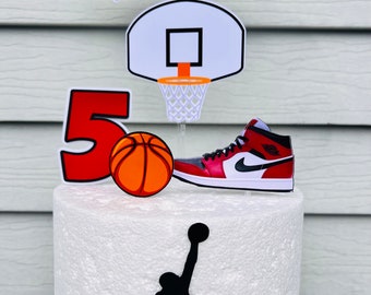 May include: A red, white, and black basketball themed cake topper with the name Mackenzie, a basketball hoop, a basketball, a red and white sneaker, and a silhouette of a basketball player.