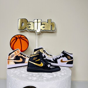 May include: A white cake decorated with a basketball, two black and gold sneakers, and a gold name topper that says "Daijah".