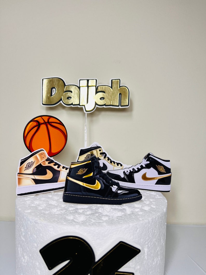 May include: A white cake decorated with a basketball, two black and gold sneakers, and a gold name topper that says "Daijah".