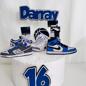 May include: A cake topper featuring a blue and white basketball theme. The topper includes cutouts of sneakers, a basketball, shoe boxes, the name "Darray", and the number "16". The sneakers are in various styles and colors.