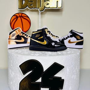 May include: A white cake with black and gold decorations. The cake is decorated with a basketball, two black and gold sneakers, the name "Daijah" and the number "24".