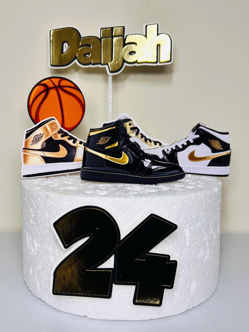 May include: A white cake with black and gold decorations. The cake is decorated with a basketball, two black and gold sneakers, the name "Daijah" and the number "24".