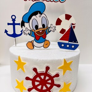 May include: A white cake decorated with a nautical theme. The cake has a blue and yellow anchor, a red and white life preserver, a blue and red sailboat, a red steering wheel, and yellow stars. The cake is topped with a figurine of a baby Donald Duck wearing a blue sailor hat and a red bow tie. The cake is also decorated with the name "Miguel".