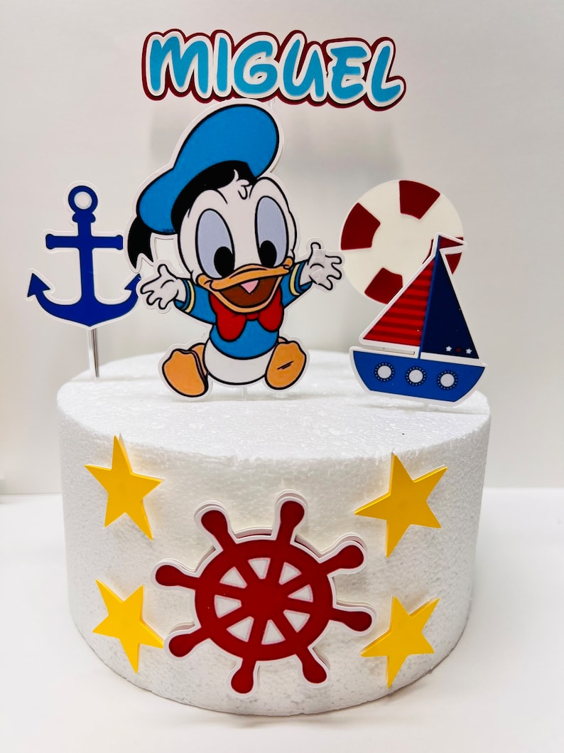 May include: A white cake decorated with a nautical theme. The cake has a blue and yellow anchor, a red and white life preserver, a blue and red sailboat, a red steering wheel, and yellow stars. The cake is topped with a figurine of a baby Donald Duck wearing a blue sailor hat and a red bow tie. The cake is also decorated with the name "Miguel".