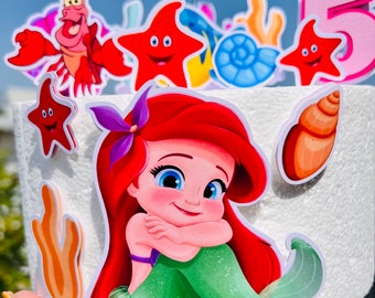 May include: A cake topper featuring a red-haired mermaid with a green tail, a purple flower in her hair, and a seashell. The topper also includes a red crab, a starfish, and a seashell.