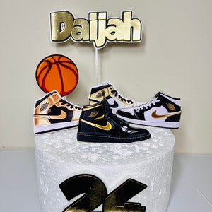 May include: A white cake decorated with a basketball, two black and gold sneakers, a gold 'Daijah' name topper, and a black and gold '24' number topper.