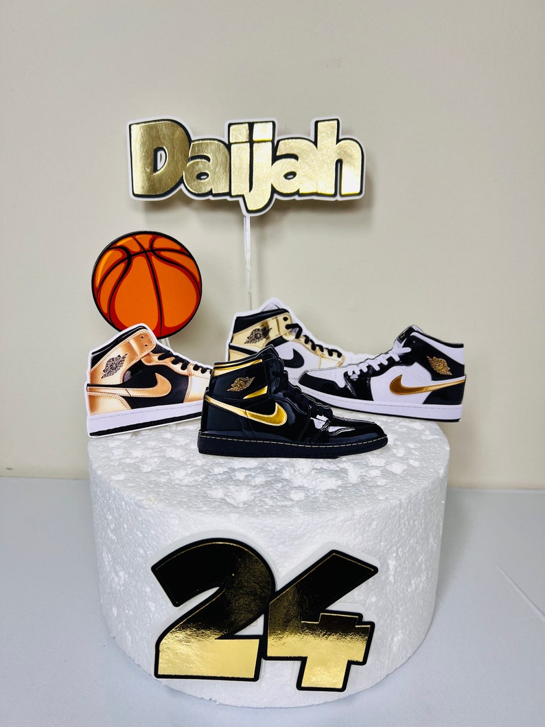 May include: A white cake decorated with a basketball, two black and gold sneakers, a gold 'Daijah' name topper, and a black and gold '24' number topper.