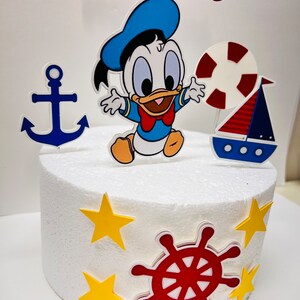 May include: A white cake decorated with a blue and yellow anchor, a red and white life preserver, a red and blue sailboat, a red steering wheel, yellow stars, and a cartoon image of a baby duck wearing a blue sailor hat. The cake is topped with a blue and white banner that says "MIGUEL".