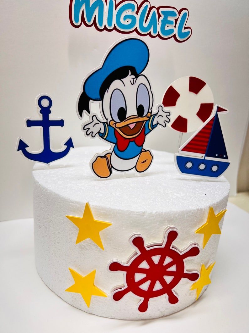 May include: A white cake decorated with a blue and yellow anchor, a red and white life preserver, a red and blue sailboat, a red steering wheel, yellow stars, and a cartoon image of a baby duck wearing a blue sailor hat. The cake is topped with a blue and white banner that says "MIGUEL".