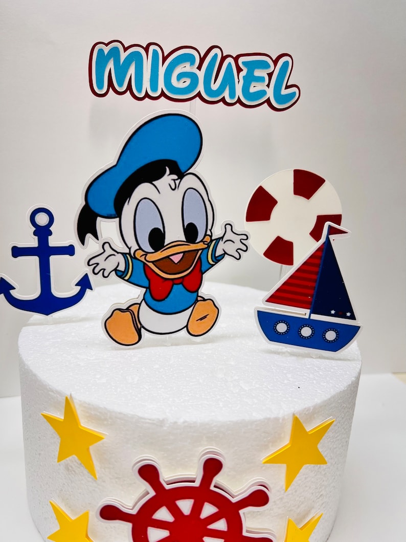 May include: A white cake decorated with a nautical theme. The cake has a blue and white baby duck character, a red and white life preserver, a blue and red sailboat, a blue anchor, a red and white steering wheel, and yellow stars. The cake is topped with a red and white banner that says "MIGUEL".