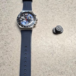May include: A blue and silver chronograph watch with a black rubber strap. The watch has a white face with black markings and a red second hand. The watch is on a white surface with a small black object to the right.