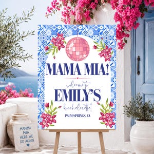 May include: A rectangular welcome sign featuring a blue and white tile pattern border, a pink disco ball, and pink floral accents. The text reads MAMA MIA! welcome to EMILY'S bachelorette PALM SPRINGS, CA. The sign is displayed on a wooden easel.