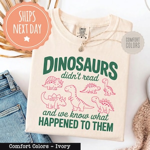 May include: Ivory-colored t-shirt with the text "DINOSAURS didn't read and we know what HAPPENED TO THEM" in green and pink. The shirt features dinosaur illustrations. The shirt is made by Comfort Colors.
