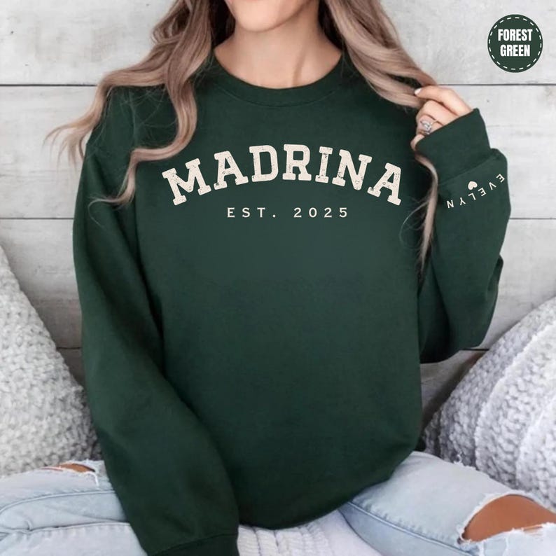 A woman wearing a green sweatshirt with the text "MADRINA EST. 2025" printed on it.