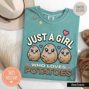 May include: A teal t-shirt featuring a graphic of three smiling cartoon potatoes with the text "JUST A GIRL WHO LOVES POTATOES" in a bold font. The design includes small pink hearts above the potatoes and decorative pink lines on the sides.