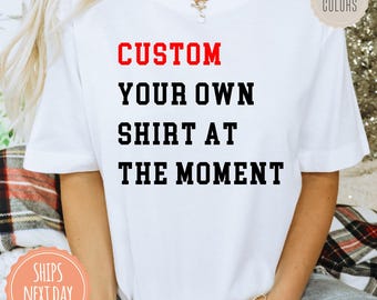 May include: A white short-sleeved t-shirt featuring the text CUSTOM in red and YOUR OWN SHIRT AT THE MOMENT in black block lettering on the front. The shirt includes a circular tag in the top right corner that reads COMFORT COLORS.