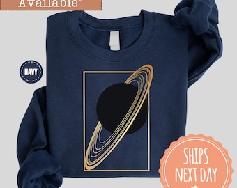 May include: Navy blue sweatshirt featuring a gold geometric design of a planet with rings inside a gold rectangle. The text "More Colors Available" is at the top, and "Navy" is in a circle on the left. The sweatshirt is folded.