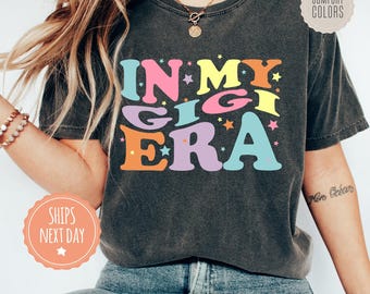 May include: A charcoal grey short-sleeve t-shirt featuring the colorful text IN MY GIGI ERA in a retro font. The design includes small multi-colored stars scattered around the lettering. The shirt has a crew neckline and a relaxed fit.