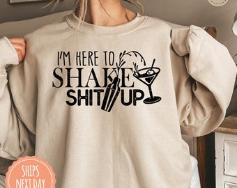May include: Sand-colored sweatshirt with the phrase "I'M HERE TO SHAKE SHIT UP" in black lettering. The graphic includes a cocktail shaker, a martini glass with an olive, and a splash effect. The sweatshirt is a casual style.