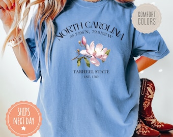 Może przedstawiać: Jasnoniebieska koszulka z nadrukiem magnolii i tekstem "NORTH CAROLINA 35.7596°N 79.0193°W TARHEEL STATE EST. 1789".