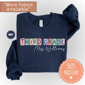 May include: Navy blue sweatshirt with the words "THIRD GRADE" in colorful block letters and the name "Mrs. Williams" in cursive. The sweatshirt has a crew neck and long sleeves. The words "More Colors Available" are at the top.
