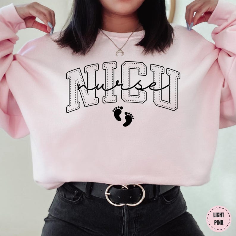 A woman wearing a pink sweatshirt with the word "NICU" printed on it, along with a pair of black pants and a black belt.
