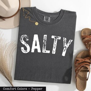 a gray t-shirt with the word "SALTY" printed on it, along with a beige hat, a pair of sandals, and some dried plants.