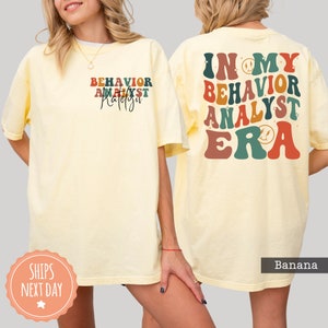 May include: A yellow t-shirt with the text "In My Behavior Analyst Era" in colorful retro font with a smiley face graphic. The text is on the back of the shirt. The front of the shirt has the text "Behavior Analyst" in a smaller font.