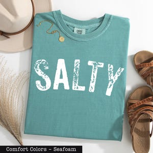 A turquoise t-shirt with the word "SALTY" printed on it, along with a beige hat, a pair of sandals, and some dried plants, all laid out on a white surface.