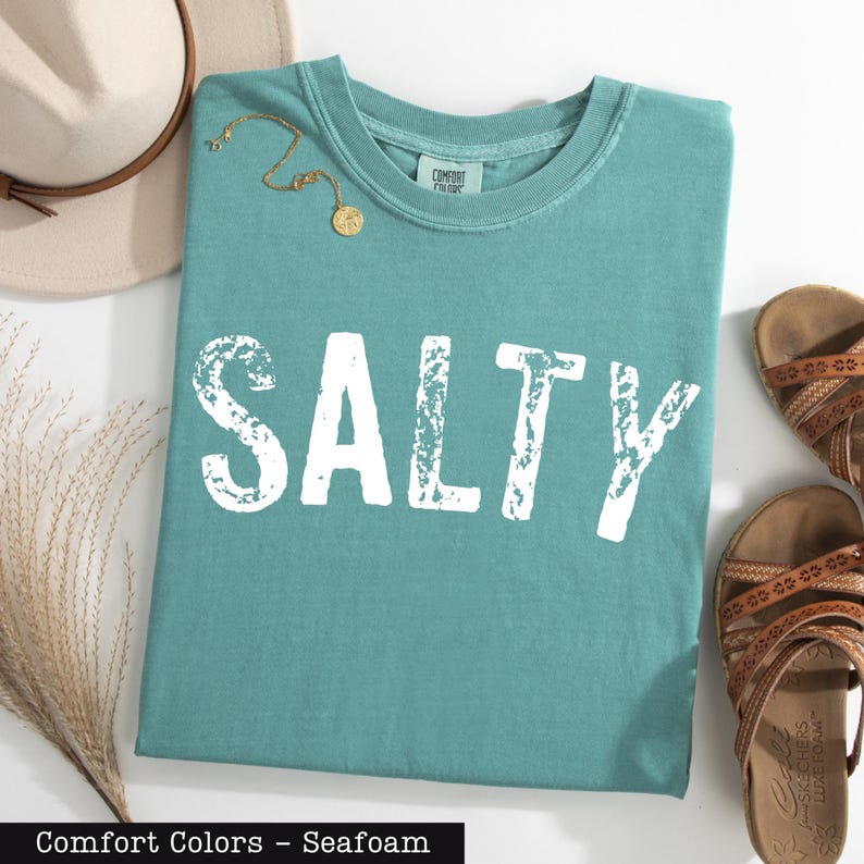 A turquoise t-shirt with the word "SALTY" printed on it, along with a beige hat, a pair of sandals, and some dried plants, all laid out on a white surface.