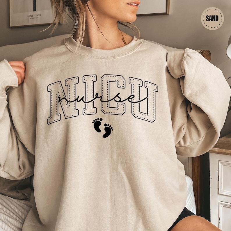 A woman wearing a beige sweatshirt with the word "NICU" printed on it, along with a small footprint symbol.
