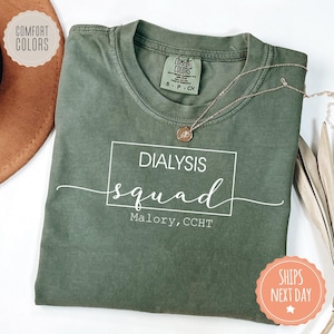 May include: Olive green t-shirt with the words "DIALYSIS squad" in white script and a box. The text "Malory, CCHT" is below the word "squad". A gold necklace is draped across the shirt. The shirt has a Comfort Colors tag.