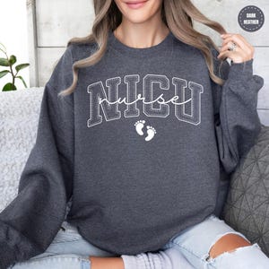 A woman wearing a grey sweatshirt with the word "NICU" printed on it, sitting on a couch.