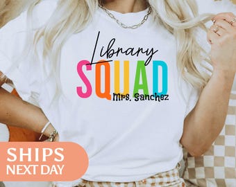 Puede incluir: Una camiseta blanca con el texto en cursiva negra Library sobre la palabra SQUAD en letras mayúsculas grandes y multicolores que incluyen rosa, naranja, amarillo, verde y azul. Debajo, el nombre Mrs. Sanchez está en negro.