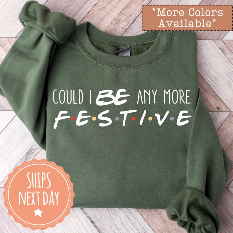 A green sweatshirt with the text "COULD I BE ANY MORE FESTIVE" printed on it, along with a "SHIPS NEXT DAY" sticker.
