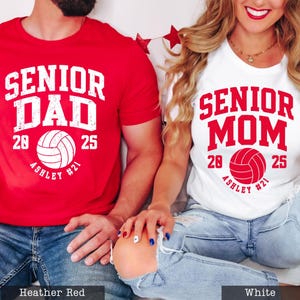 A man and a woman wearing matching t-shirts with the text "SENIOR DAD" and "SENIOR MOM" printed on them, along with the year "2025" and the volleyball team name "ASHLEY #21".