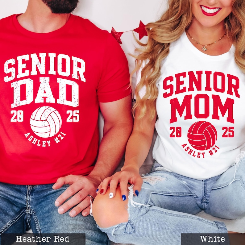 A man and a woman wearing matching t-shirts with the text "SENIOR DAD" and "SENIOR MOM" printed on them, along with the year "2025" and the volleyball team name "ASHLEY #21".