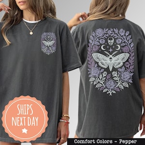 May include: Dark grey t-shirt with a moth and floral design on the front and back. The back features a moth, skull, crescent moons, and roses. The front has a smaller moth and floral design. The text "Comfort Colors - Pepper" is at the bottom.