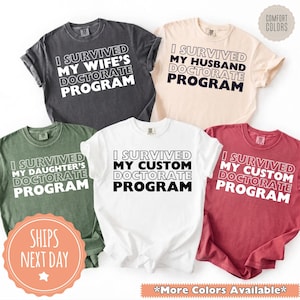 May include: A collection of Comfort Colors t-shirts in various colors, each with the phrase "I SURVIVED MY [WIFE'S/HUSBAND'S/DAUGHTER'S/CUSTOM] DOCTORATE PROGRAM" printed in bold white or black letters. The shirts are displayed on a white background.