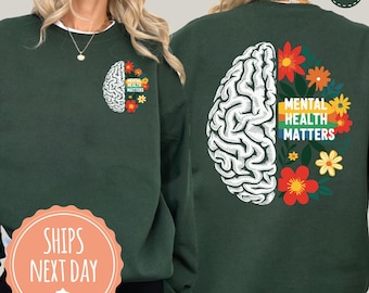 Puede incluir: Sudadera verde bosque con un gráfico de cerebro blanco y el texto "Mental Health Matters" rodeado de flores coloridas. El diseño se repite en la parte delantera y trasera de la sudadera.