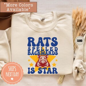 May include: Cream-colored sweatshirt with the text "RATS SPELLED BACKWARDS IS STAR" in blue and yellow. A cartoon rat is centered on a yellow star. The sweatshirt is a casual style.