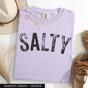 a purple t-shirt with the word "SALTY" printed on it, along with a beige hat, a pair of sandals, and some dried flowers.