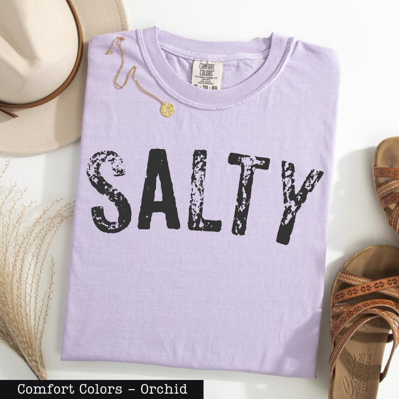 a purple t-shirt with the word "SALTY" printed on it, along with a beige hat, a pair of sandals, and some dried flowers.