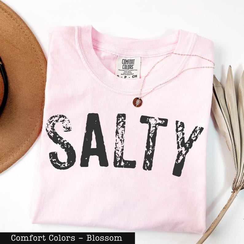 A pink t-shirt with the word "SALTY" printed on it, along with a brown hat and some dried leaves, all placed on a white surface.