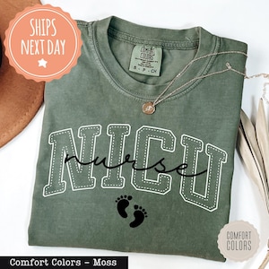 May include: A moss green Comfort Colors t-shirt with the text "NICU nurse" in white, featuring a collegiate font and a cursive "nurse" script. Below the text are two black baby footprints. A gold necklace rests on the shirt.