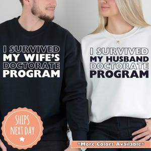 May include: A black sweatshirt with the text "I SURVIVED MY WIFE'S DOCTORATE PROGRAM" and a white sweatshirt with the text "I SURVIVED MY HUSBAND DOCTORATE PROGRAM". The sweatshirts are displayed side by side.