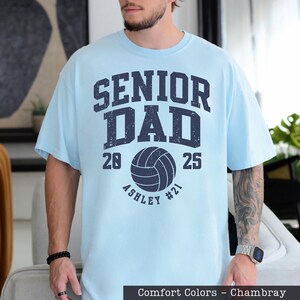 A man wearing a light blue t-shirt with a graphic design that says "SENIOR DAD" and "ASHLEY #21" printed on it.